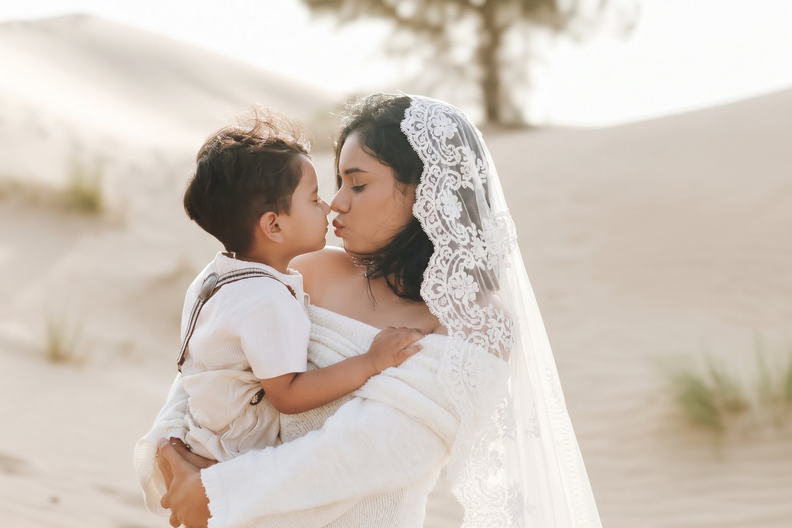 Bride in a lace veil kissing her son in her arms in the desert