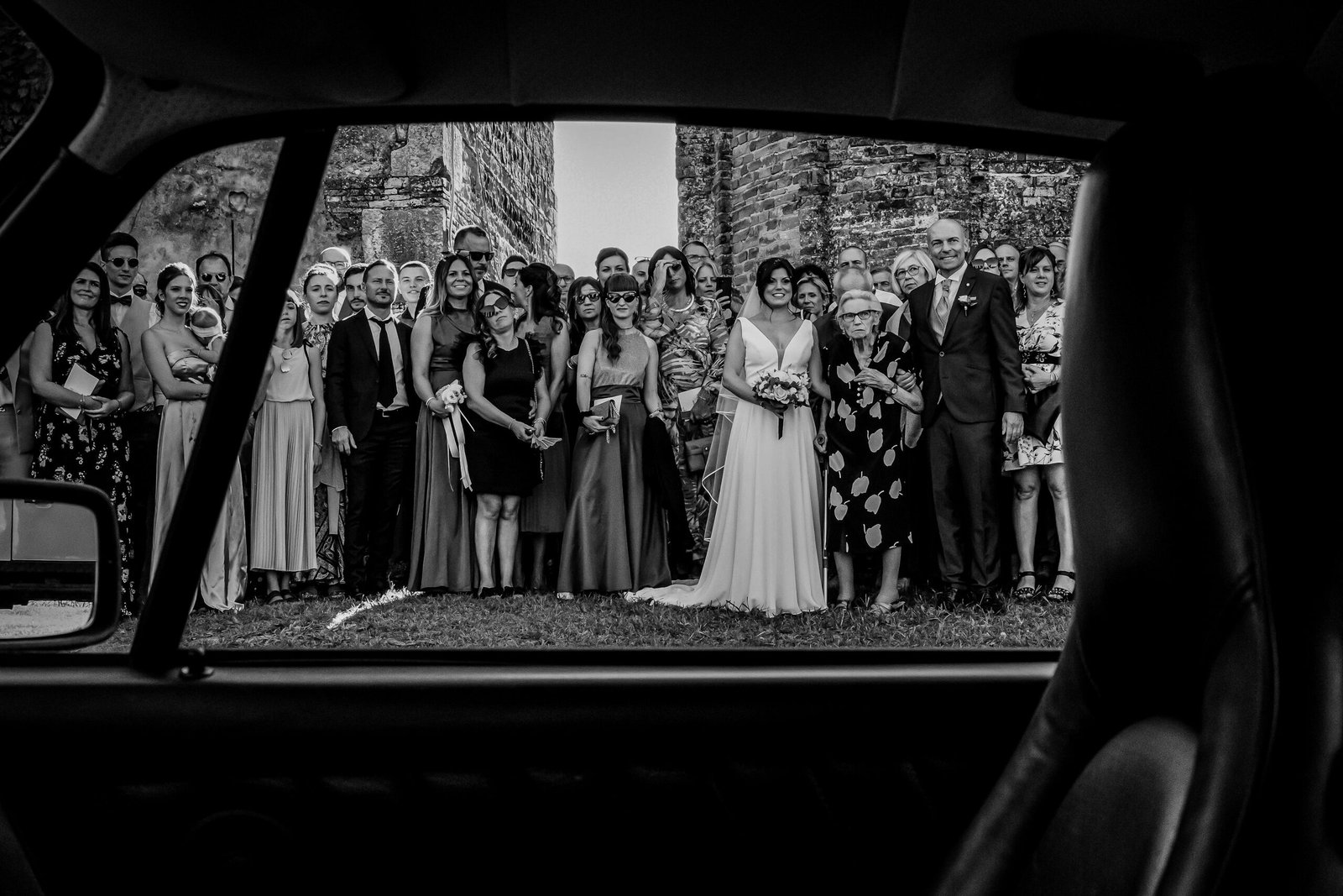 Sposi e invitati fotografati attraverso il finestrino di un’auto d’epoca, in bianco e nero