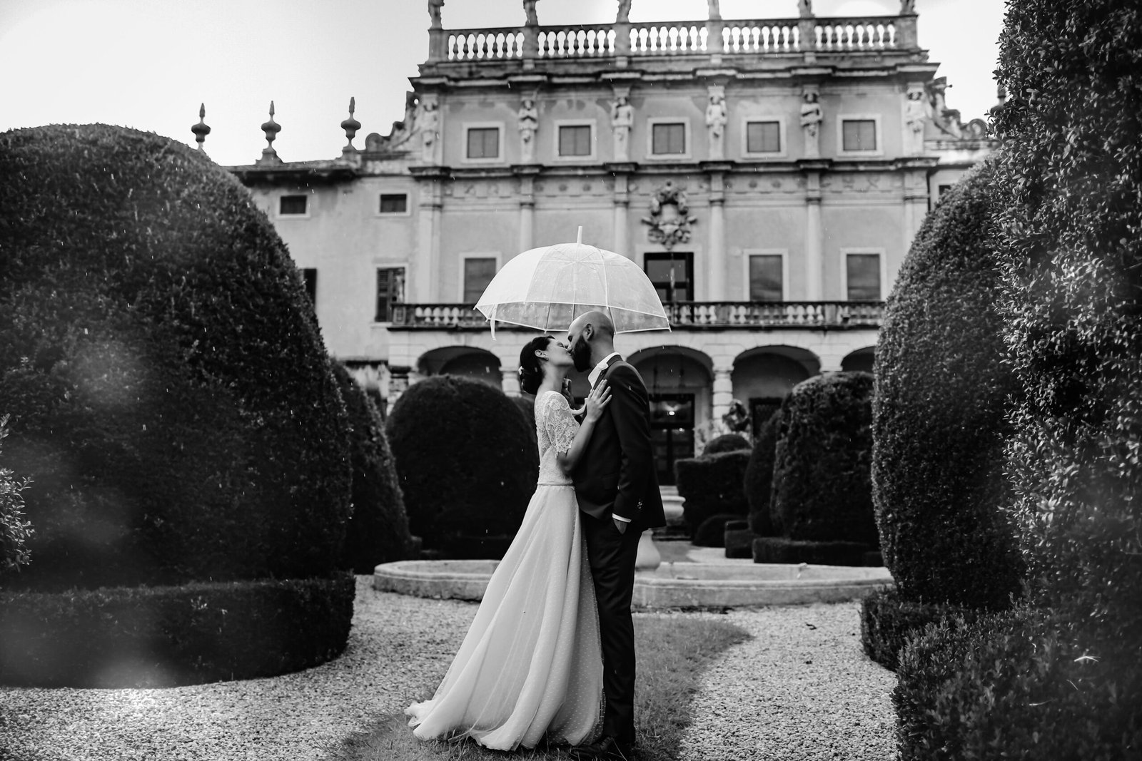 Sposi sotto un ombrello trasparente nel giardino di una villa storica, in bianco e nero