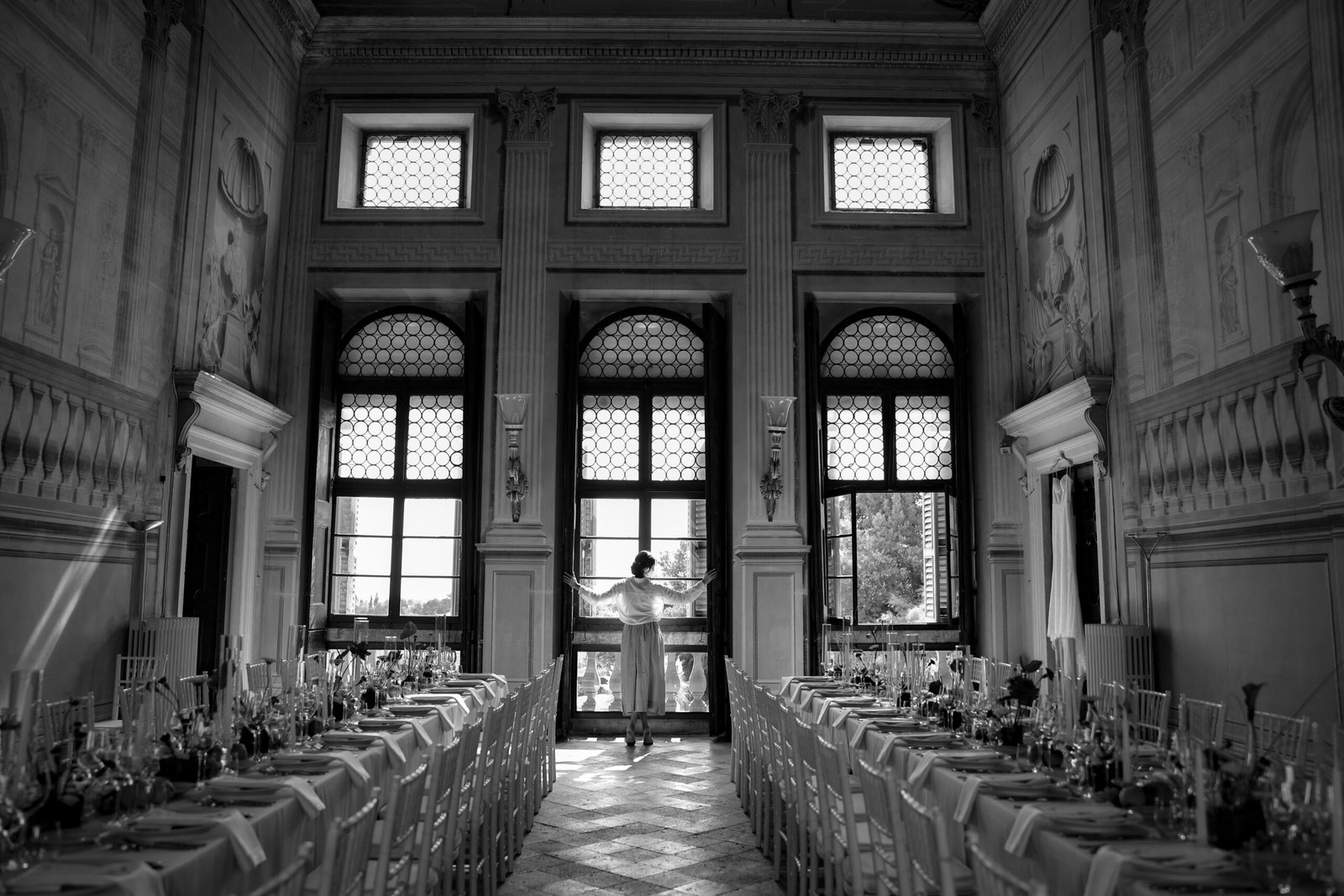 Bride with arms open in front of the windows of the hall set up for the reception, in black and white