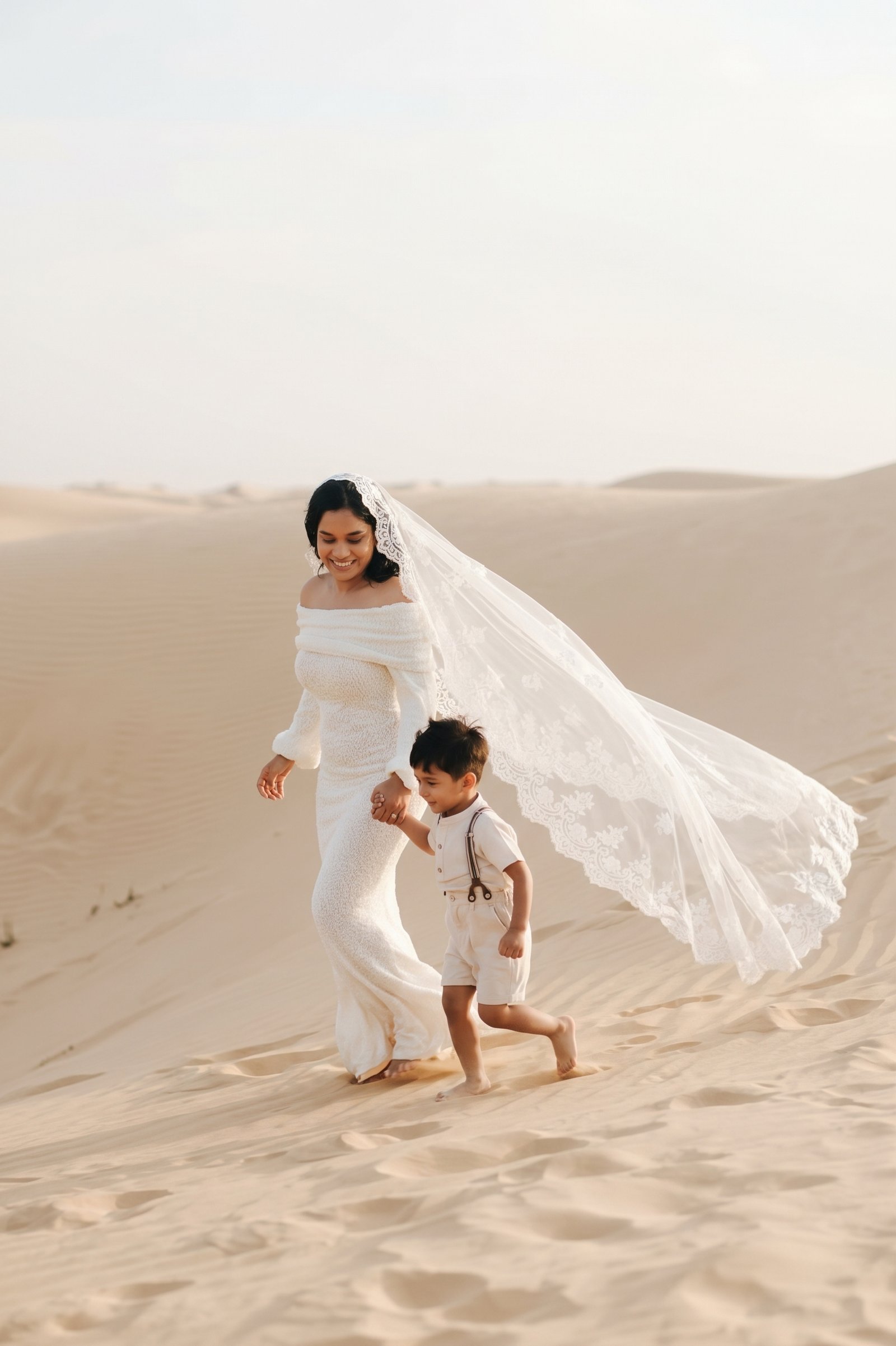 Bride with veil blowing in the wind walking hand in hand with her son through the dunes of the Dubai desert