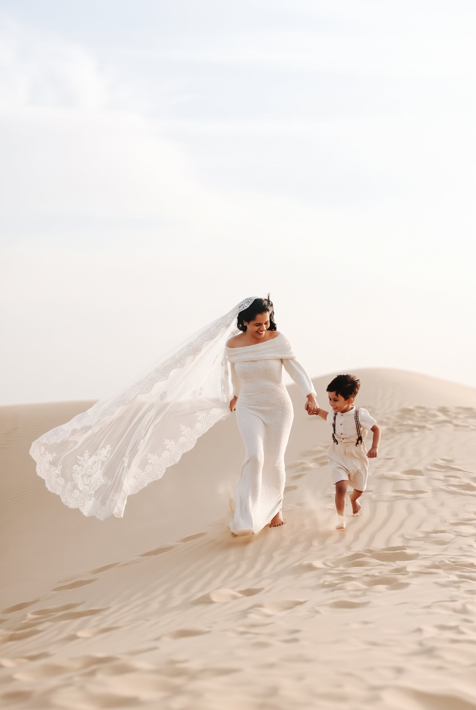 Bride and son running across the desert dunes at sunset, her veil flying in the wind