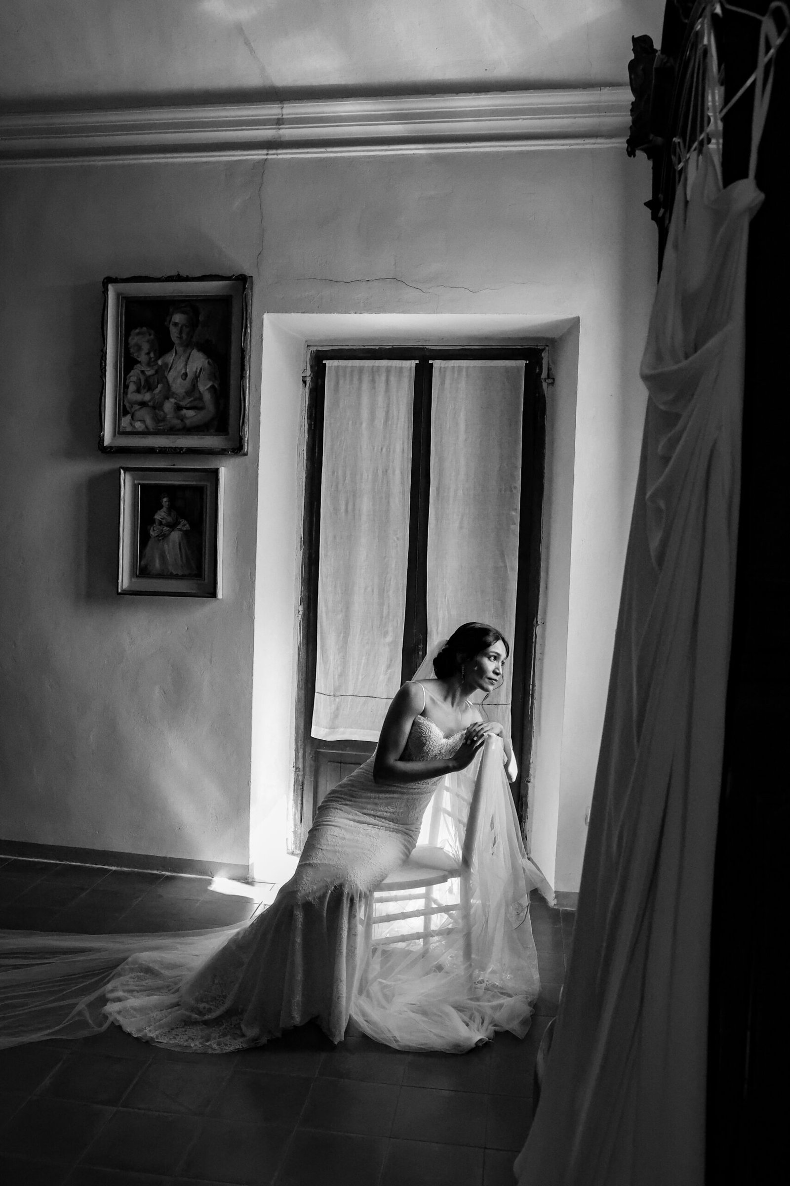 Bride seated by the window, adjusting her dress, in black and white