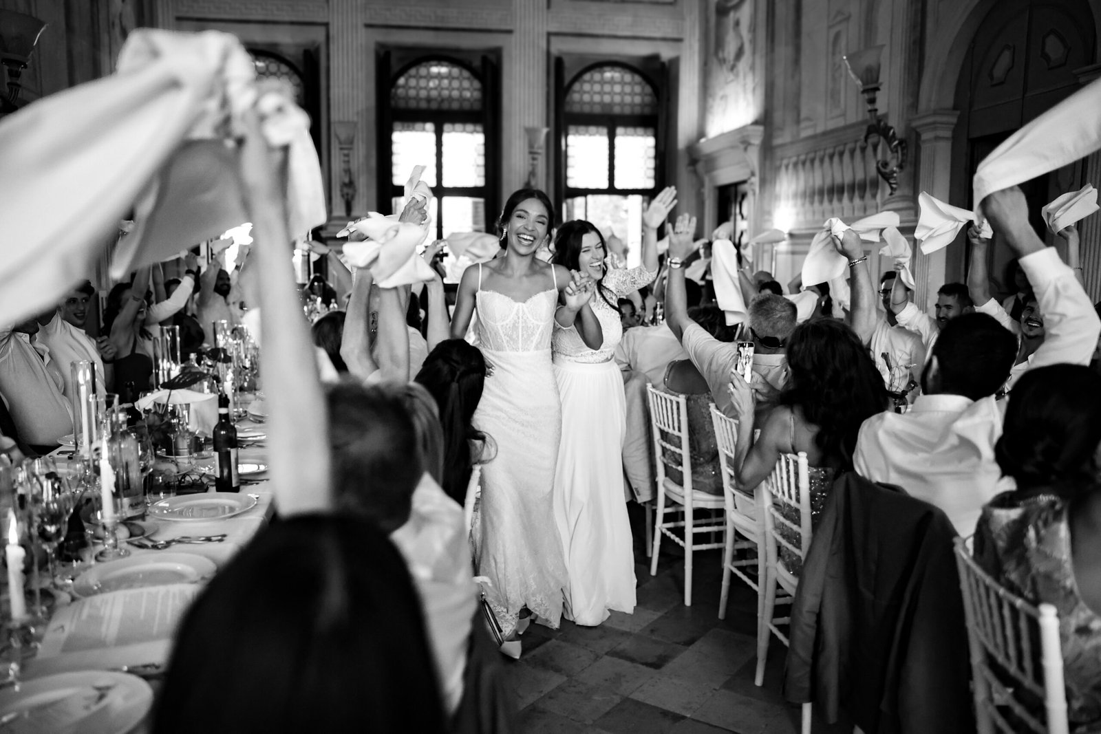 Two brides entering the reception hall of Villa Fraccaroli, welcomed by guests waving napkins, in black and white
