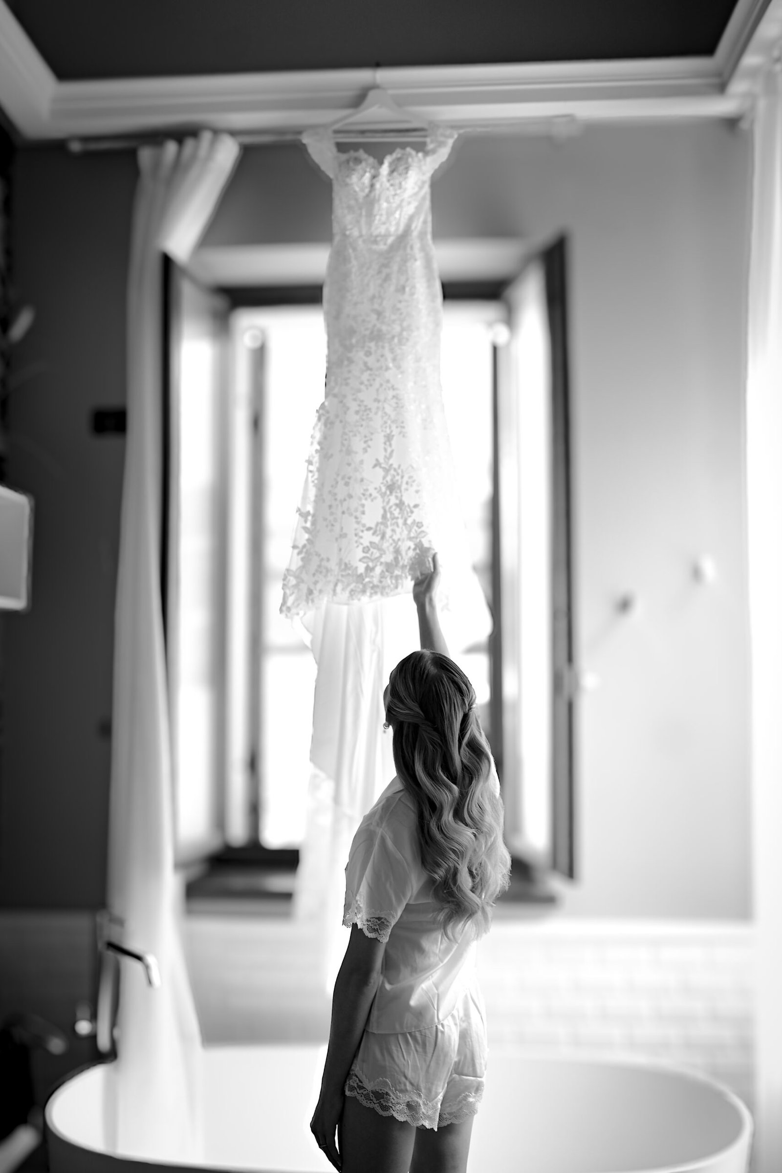 Bride backlit, brushing her hand against the dress hanging in the window during the preparations, in black and white