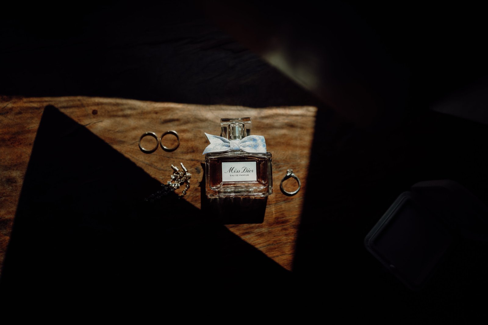 Dettagli della sposa: fedi, orecchini e profumo su un tavolo in legno illuminato da un raggio di luce