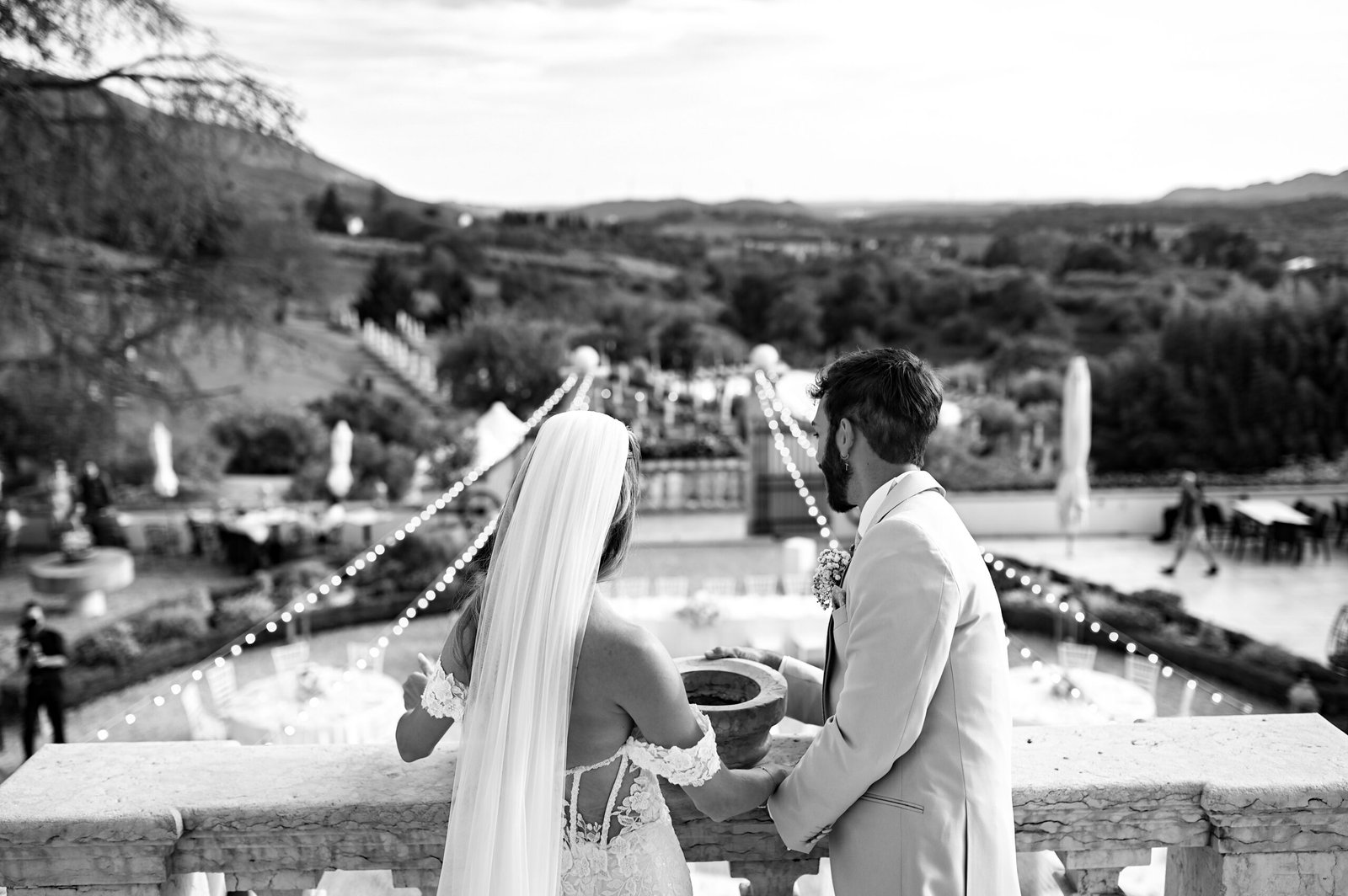 Bride and groom at the terrace balustrade overlooking the hills and the reception, in black and white