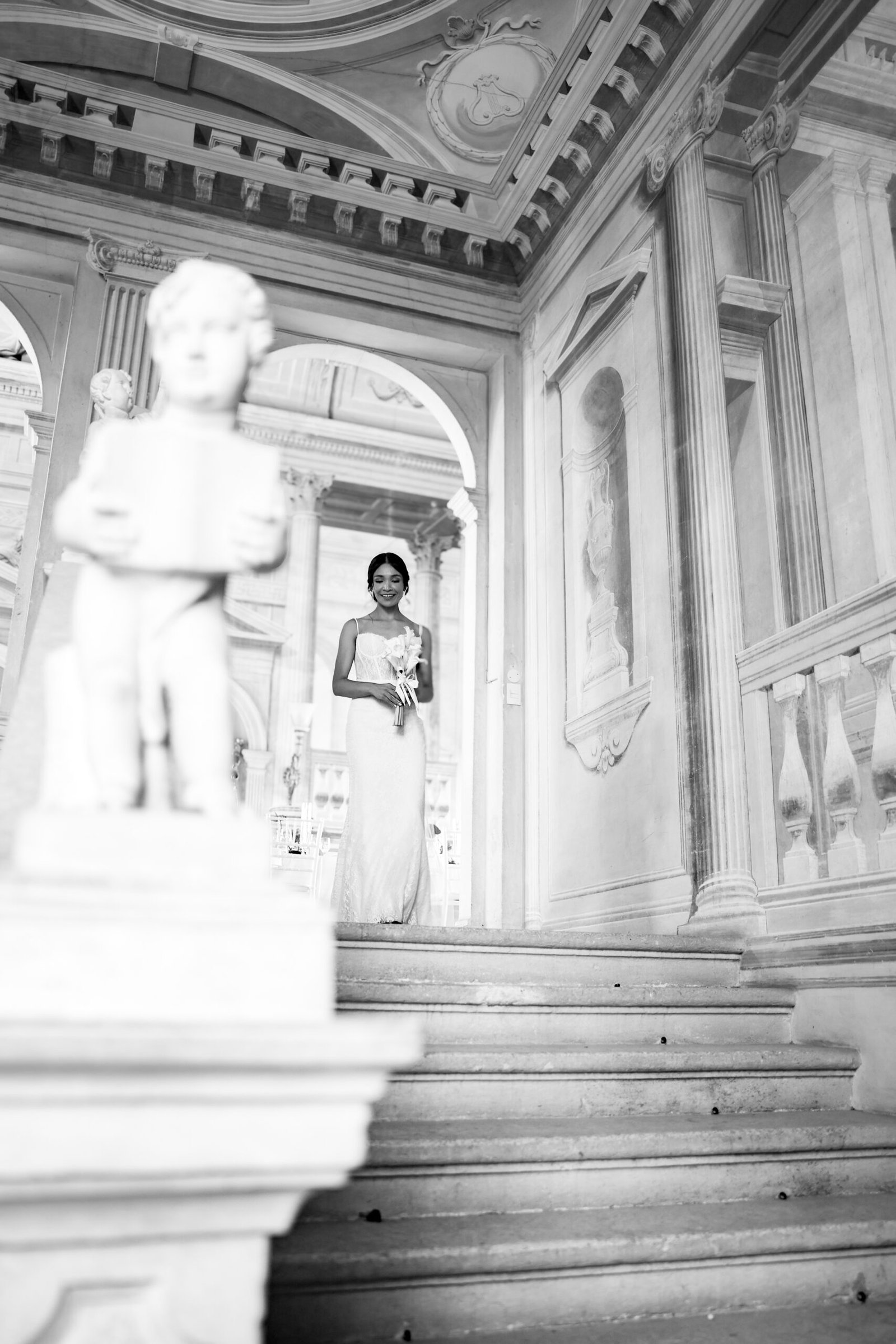 Bride at the top of the frescoed staircase of Villa Fraccaroli, next to a statue, in black and white